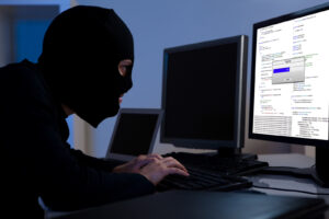 Masked hacker wearing a balaclava sitting at a desk downloading private information off a computer. Avoiding fraud.